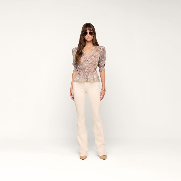 A woman wearing sunglasses, the Suri Top, cream-colored flared pants, and beige shoes stands against a plain white background.