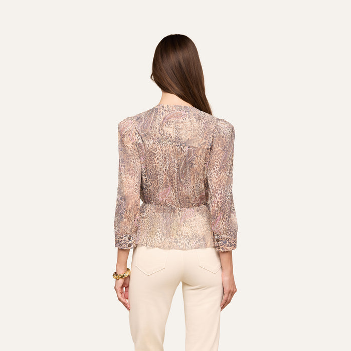 A woman with long brown hair, wearing the Suri Top—a sheer paisley animal print blouse with three-quarter sleeves—and cream-colored pants stands facing away from the camera against a plain background.