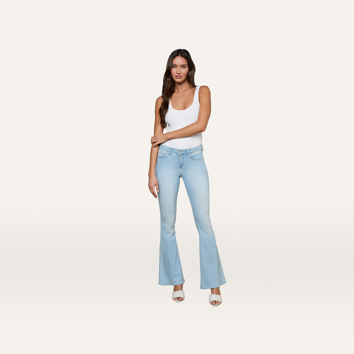 A woman stands against a plain white background, facing forward with one arm crossed, wearing a white tank top and the Taney Low-Rise Flare Jean in light blue.