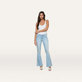 A woman stands against a plain white background, facing forward with one arm crossed, wearing a white tank top and the Taney Low-Rise Flare Jean in light blue.