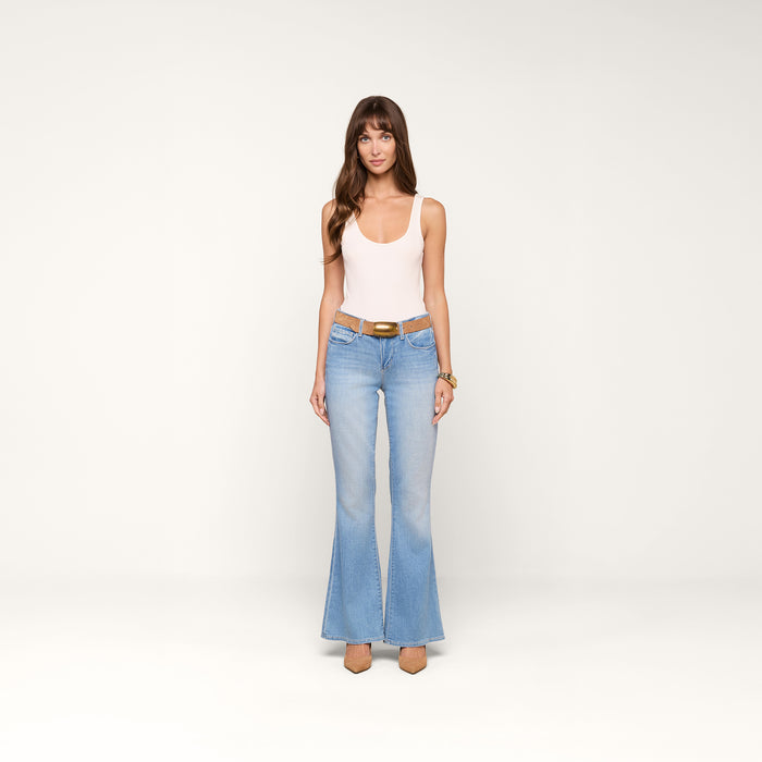A woman stands against a plain white background, wearing a light pink tank top, the Taney Low-Rise Flare Jean in a light indigo wash with a brown belt, nude heels, and gold bracelets.