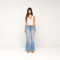 A woman stands against a plain white background, wearing a light pink tank top, the Taney Low-Rise Flare Jean in a light indigo wash with a brown belt, nude heels, and gold bracelets.