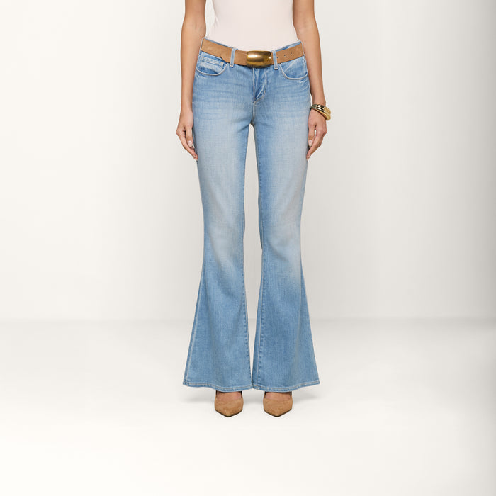A person wearing the Taney Low-Rise Flare Jean in a light indigo wash, paired with a tan belt, white top, and brown pointed-toe shoes, stands against a plain white background with only their torso and legs visible.