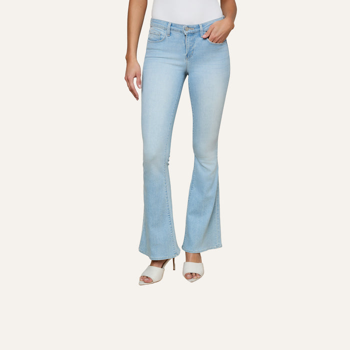 A person wearing Taney Low-Rise Flare Jeans in light blue stretch denim and white heeled sandals stands against a plain background, with only the lower half of the body visible.