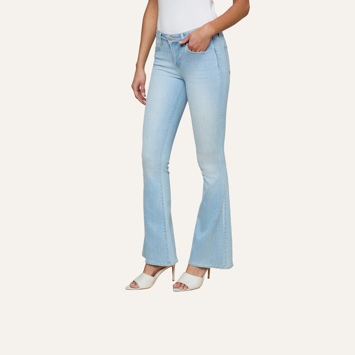 Wearing the Taney Low-Rise Flare Jean in light blue stretch denim, a person stands with one hand in their pocket, paired with a white sleeveless top and beige heeled sandals against a plain background.