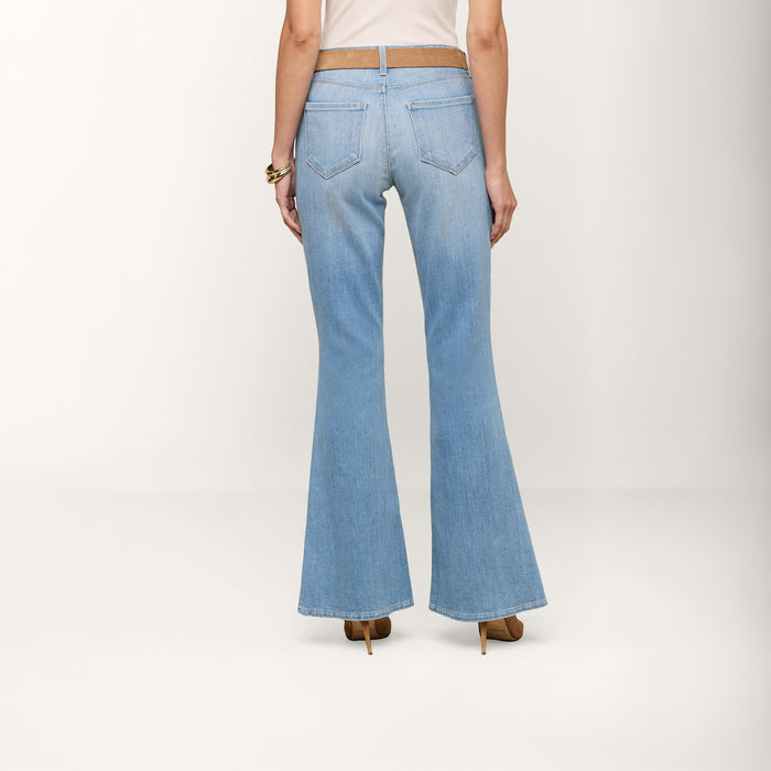 A person is shown from the back, standing against a plain white background, wearing the Taney Low-Rise Flare Jean in a light indigo wash paired with brown heels.