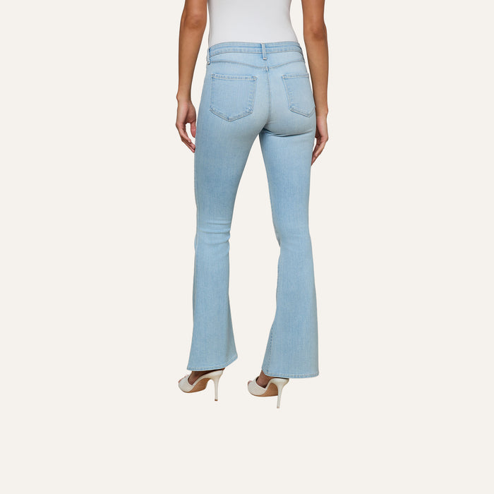 A woman stands facing away against a plain light background, showcasing the back of the Taney Low-Rise Flare Jean in light blue stretch denim, paired with white heels.