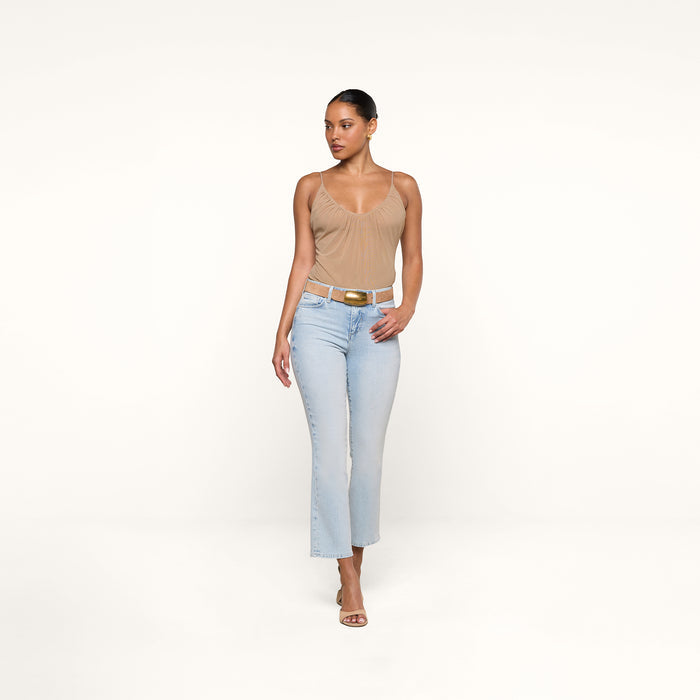 A woman wearing a beige sleeveless top, the Tati Cropped Micro Boot Jean in a light wash stretch denim, a white belt, and heeled sandals stands against a plain white background.