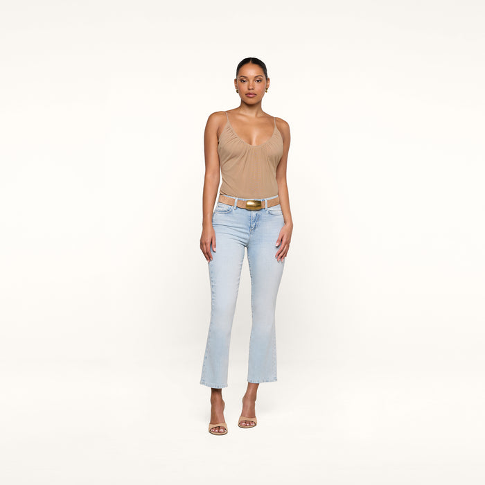 A woman stands against a plain white background, wearing a tan sleeveless top, the Tati Cropped Micro Boot Jean in light wash stretch denim with a cropped kick flare, a white belt with a gold buckle, and tan high-heeled sandals. She faces forward with her arms relaxed at her sides.