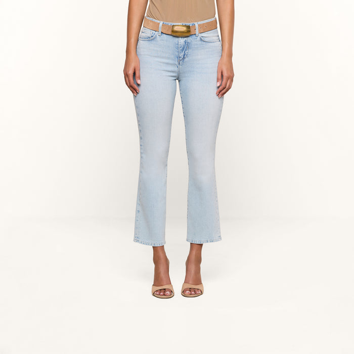 A person wearing the Tati Cropped Micro Boot Jean in a light wash, paired with a beige top, a brown belt, and beige heeled sandals stands against a plain white background.