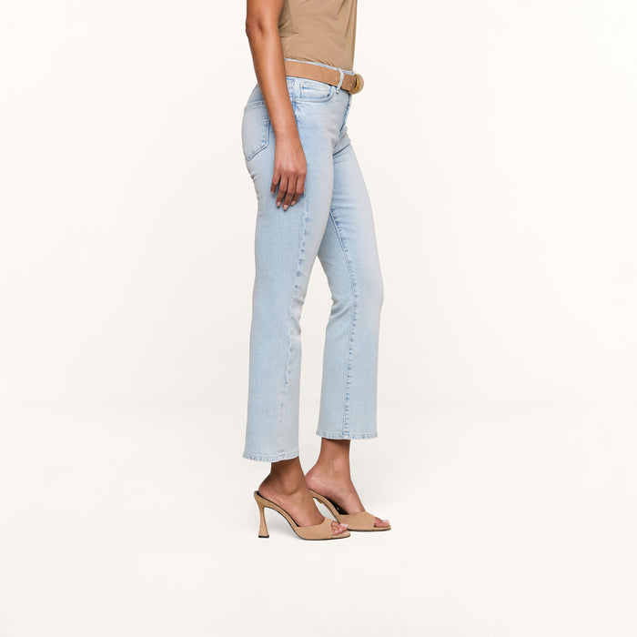 A person stands sideways against a plain white background, wearing Tati Cropped Micro Boot Jeans in a light wash stretch denim, styled with a beige belt, tan top, and beige heeled sandals.