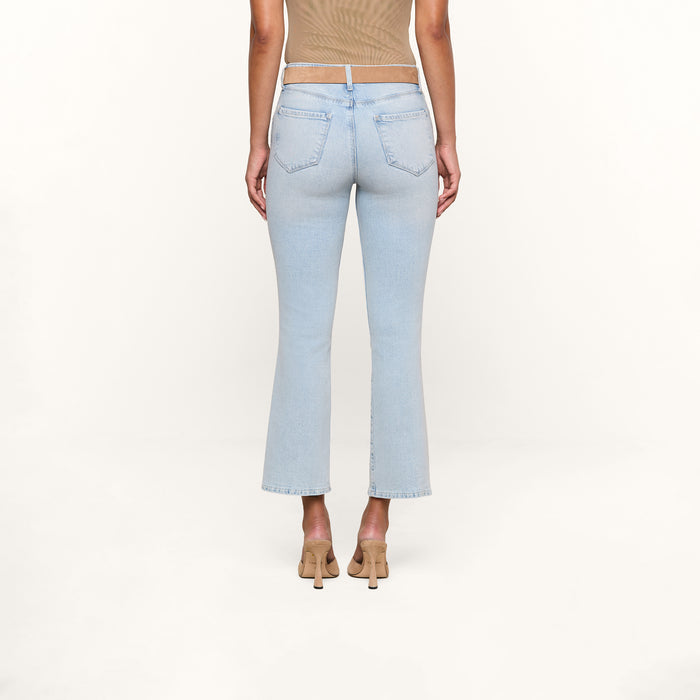A person stands facing away, wearing the Tati Cropped Micro Boot Jean in a light wash, paired with a taupe sleeveless top and beige heeled shoes against a plain white background.