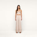 A woman stands facing forward wearing a light beige crop top and the Tavia Trouser — a high-waisted, wide-leg satin pant with dark trim at the hem — paired with open-toe shoes. The background is plain white.