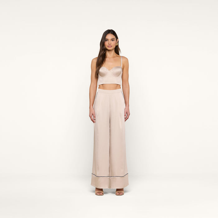 A woman stands facing forward wearing a light beige crop top and the Tavia Trouser — a high-waisted, wide-leg satin pant with dark trim at the hem — paired with open-toe shoes. The background is plain white.