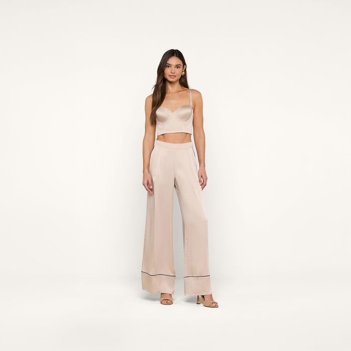 A woman stands against a plain white background wearing a light beige camisole and the Tavia Trouser in matching satin with dark trim at the hems. She has long brown hair and is wearing open-toe heels, capturing the effortless elegance of luxe loungewear.