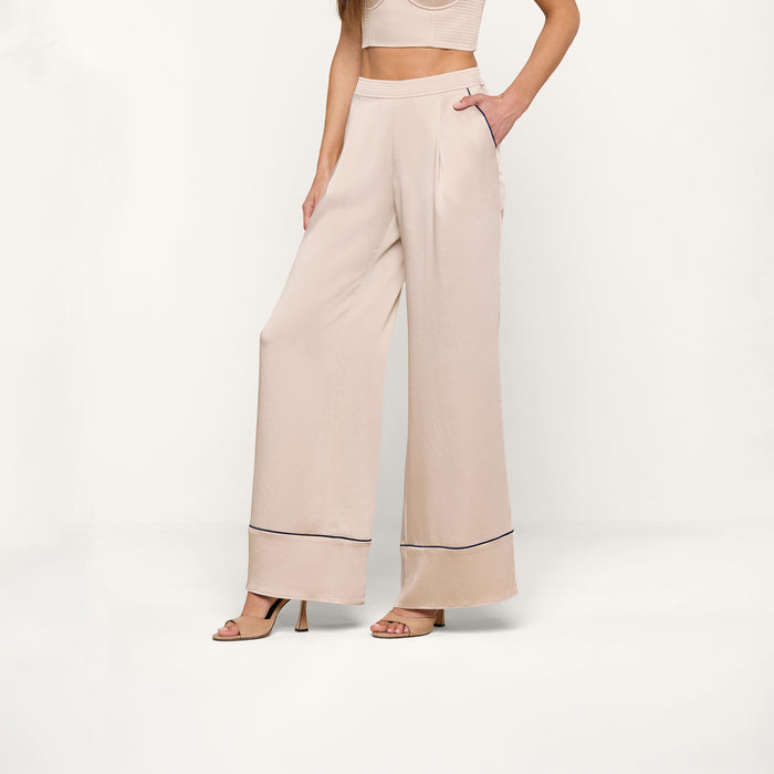 A woman stands against a white background, wearing the high-waisted, wide-leg Tavia Trouser in beige satin featuring black trim near the hem. She pairs it with a matching crop top and nude heeled sandals. One hand rests in her pocket, and her face is not visible.