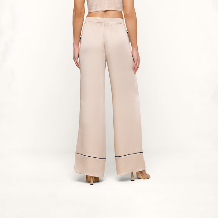 A woman stands facing away, wearing the Tavia Trouser—high-waisted, wide-leg satin trousers with a thin dark trim near the hem—paired with beige heeled sandals and a cropped beige top.