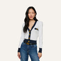 Wearing the Taya Cardigan—a Parisian-inspired white layer detailed with metallic trim, gold buttons, and a chest pocket—paired with blue jeans, the woman stands against a plain light background.
