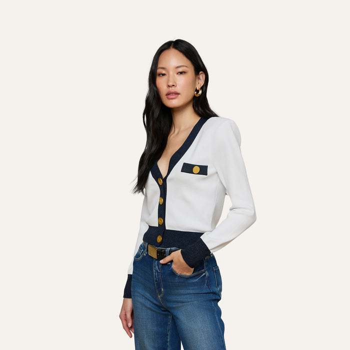 A woman with long black hair wears the Taya Cardigan, a Parisian-inspired white piece featuring navy and metallic trim with gold buttons, paired with blue jeans. She stands with one hand in her pocket against a plain light background.