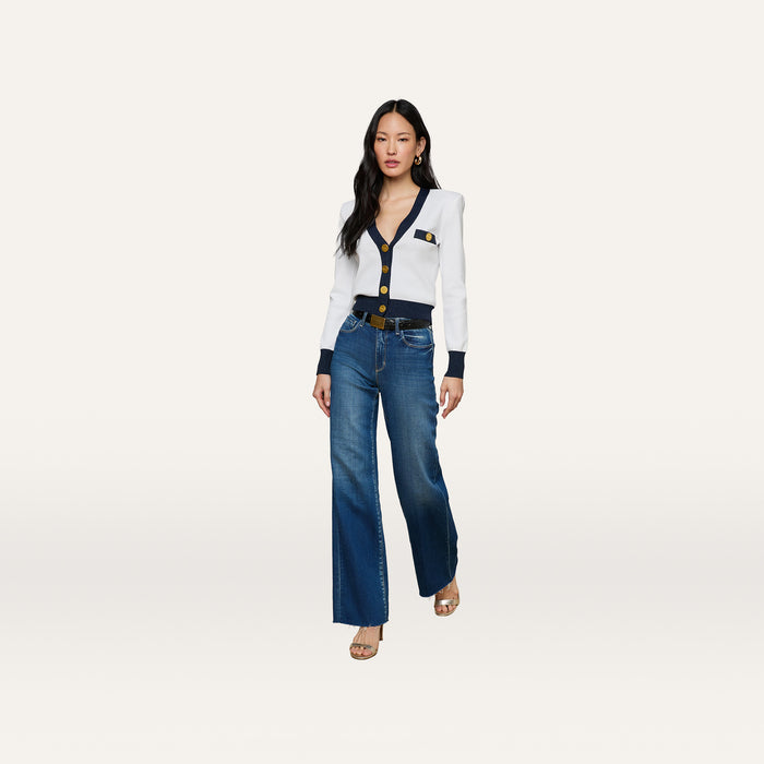 A woman stands facing forward, wearing the Taya Cardigan—a Parisian-inspired white cardigan with navy and metallic trim and gold buttons—paired with blue wide-leg jeans, a black belt, and heeled sandals against a plain light background.