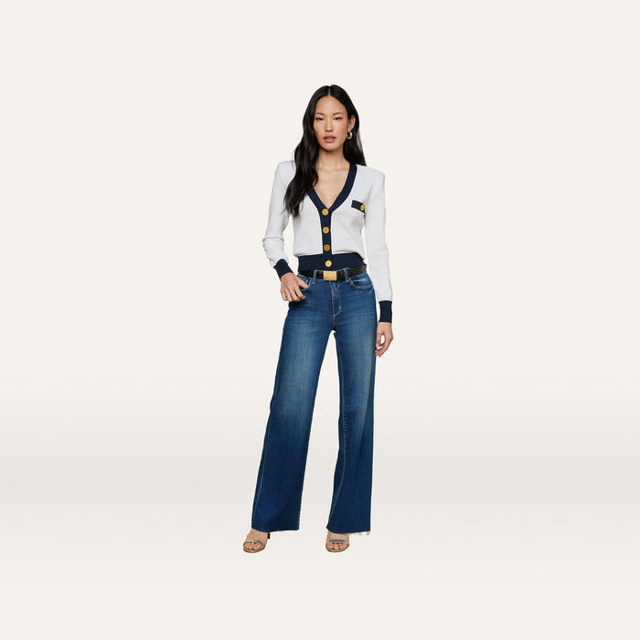 A woman with long dark hair and a neutral expression stands against a white background, wearing the Taya Cardigan—a Parisian-inspired white and navy piece featuring gold buttons and metallic trim—paired with blue wide-leg jeans and beige heels.