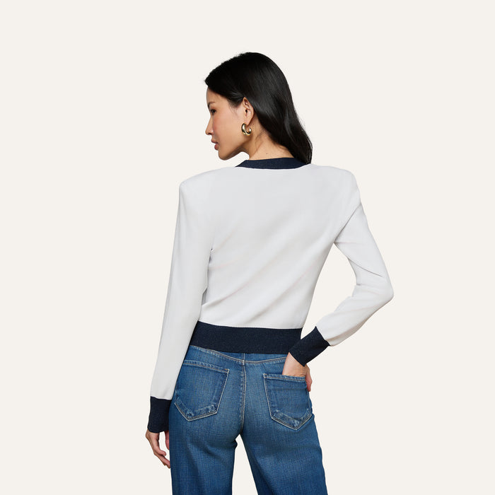 A woman with long dark hair stands with her back to the camera, wearing the Parisian-inspired Taya Cardigan in white with dark cuffs and a metallic trim, paired with blue jeans. She rests her right hand in her back pocket.