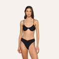 A woman stands facing forward, wearing the Tenali Bikini Top, an underwire black bra bikini top paired with high-waisted, V-cut bottoms. The background is plain and light-colored.