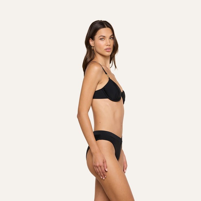 A woman stands in profile against a plain, light-colored background, wearing the Tenali Bikini Top in black. She has long brown hair, a neutral expression, and minimal jewelry.