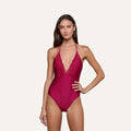 Wearing the Terra One-Piece Swimsuit, a woman poses against a plain light background; the suit features a deep red hue, lace-trimmed details, a plunging V neckline, and halter straps.