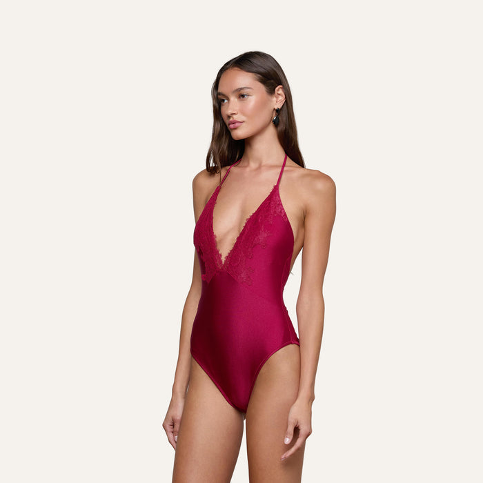 A woman stands against a plain light background wearing the Terra One-Piece Swimsuit in deep red, featuring lace trim and a deep V neckline.
