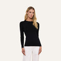 A person with long blonde hair wears the fitted black Tess Long Sleeve Tee and white pants, standing against a plain light background.
