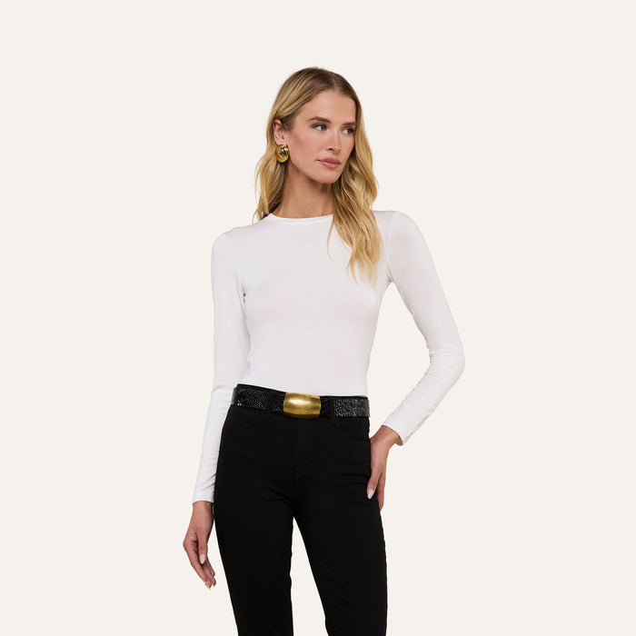 A woman with long blonde hair wears the Tess Long Sleeve Tee in white, paired with black pants, a wide black belt featuring a large gold buckle, and gold earrings. She stands against a plain light background, glancing slightly to the side.