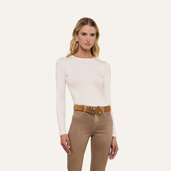 Woman with long blonde hair wearing the Tess Long Sleeve Tee in white, styled with tan pants and a brown belt, standing against a plain light background.