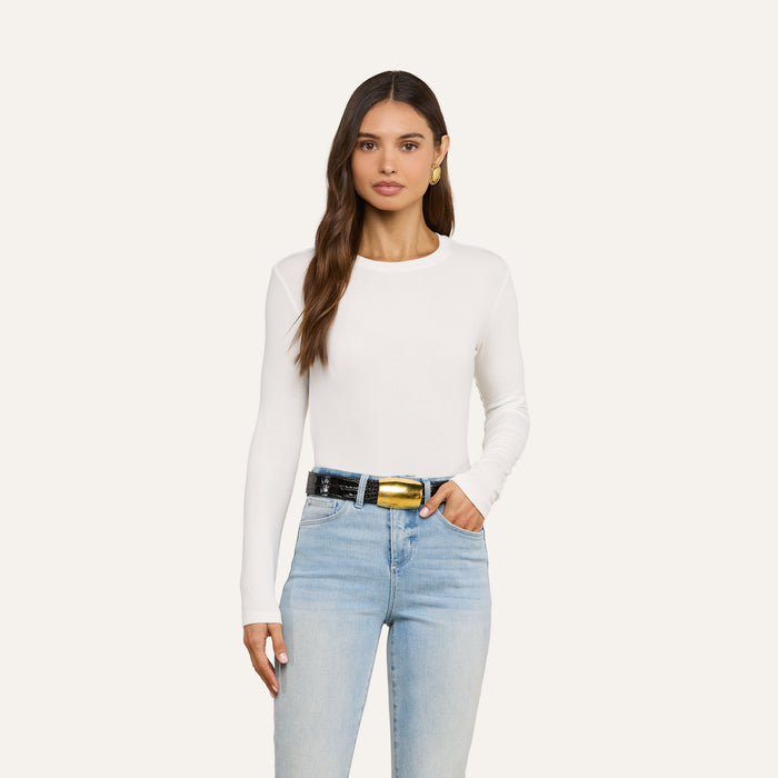 A woman with long brown hair stands against an off-white background wearing the Tess Long Sleeve Tee, a slim-fit white crew neck top, paired with light blue jeans, a black belt featuring a large gold buckle, and gold hoop earrings.