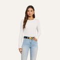 A woman with long brown hair stands against an off-white background wearing the Tess Long Sleeve Tee, a slim-fit white crew neck top, paired with light blue jeans, a black belt featuring a large gold buckle, and gold hoop earrings.