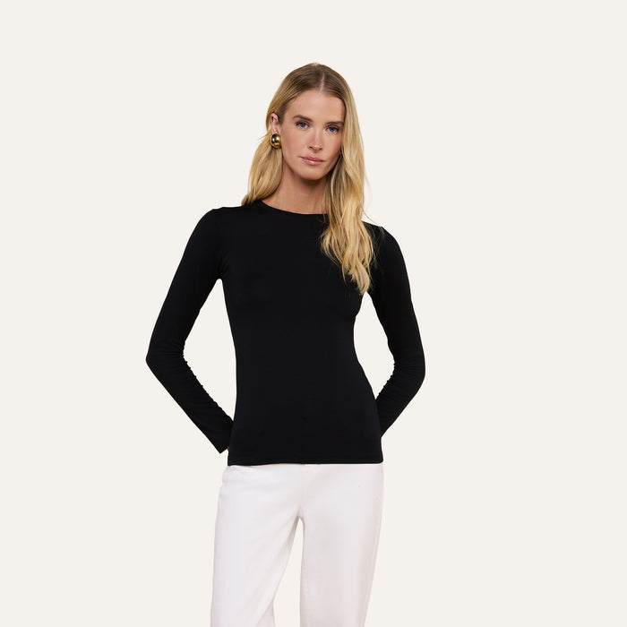 A woman with long blonde hair wears the fitted black Tess Long Sleeve Tee and white pants, standing against a plain light background with her hands behind her back.