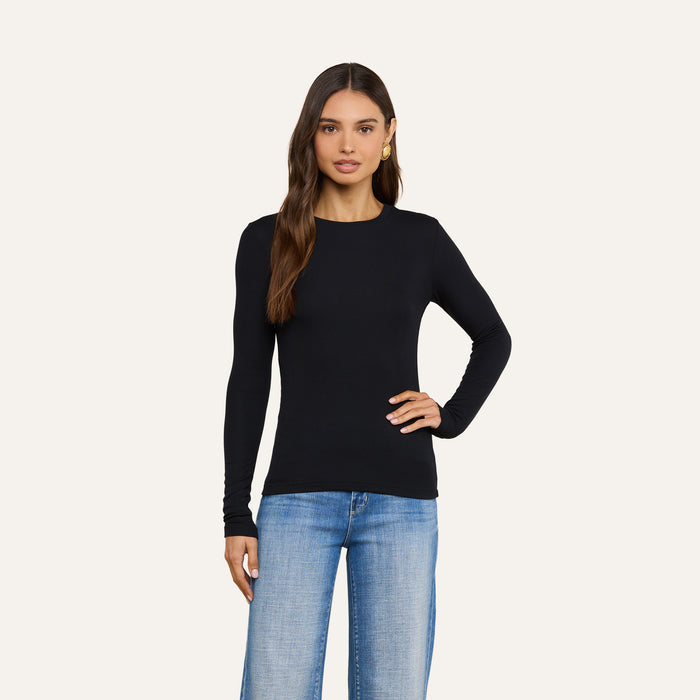 A woman with long brown hair wearing the Tess Long Sleeve Tee in black, paired with light blue jeans, stands with one hand on her hip against a plain light background.