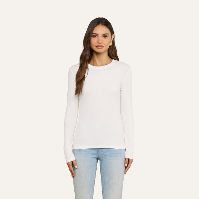 Wearing the Tess Long Sleeve Tee in white, a woman with long brown hair and a neutral expression stands facing forward against a light background, paired with light blue jeans.