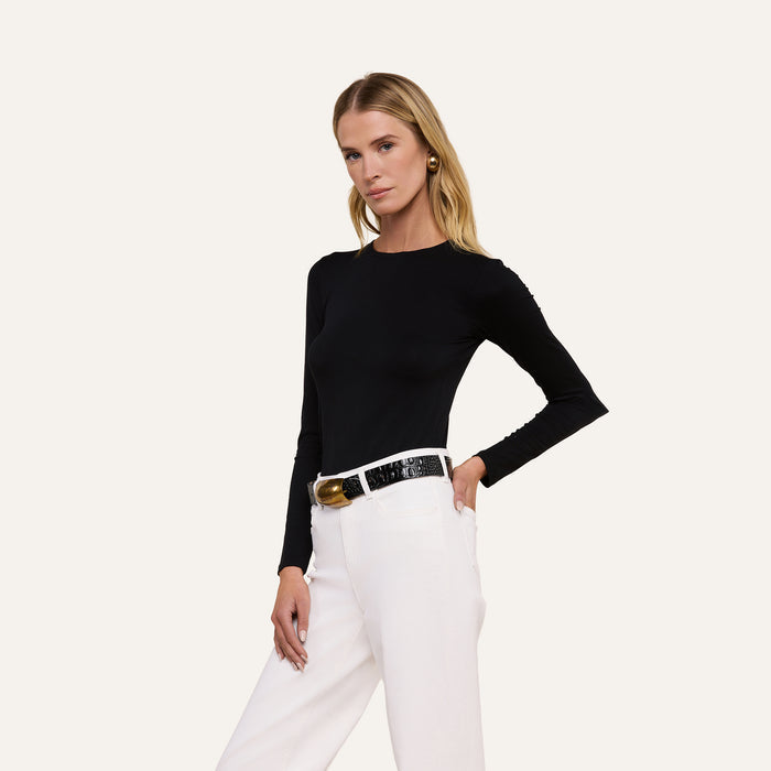 A woman stands confidently against a plain light background, wearing the Tess Long Sleeve Tee in black with white high-waisted pants and a black belt, looking directly at the camera.