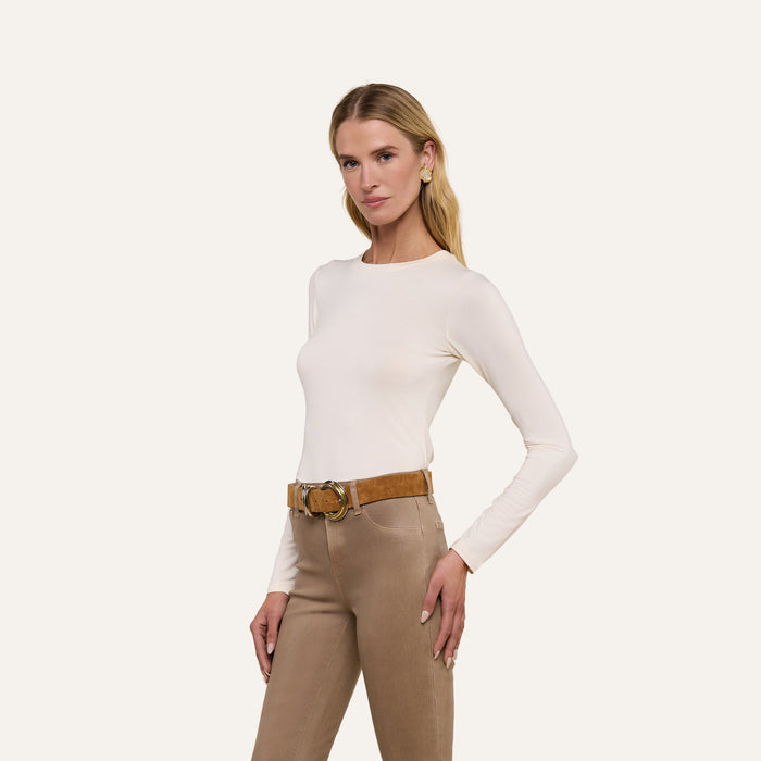 A woman poses with one hand on her hip against a plain light background, wearing the Tess Long Sleeve Tee in white, tan pants, and a wide brown belt.