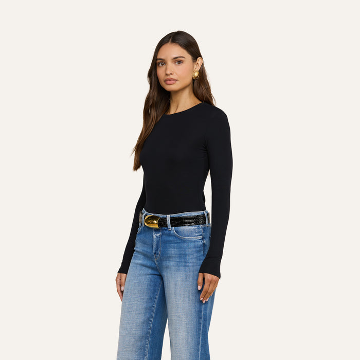 A woman with long brown hair wears the Tess Long Sleeve Tee—a slim-fit black long sleeve top—paired with blue jeans, a black belt with a gold buckle, and gold earrings. She stands against a plain light background, looking slightly to the side.