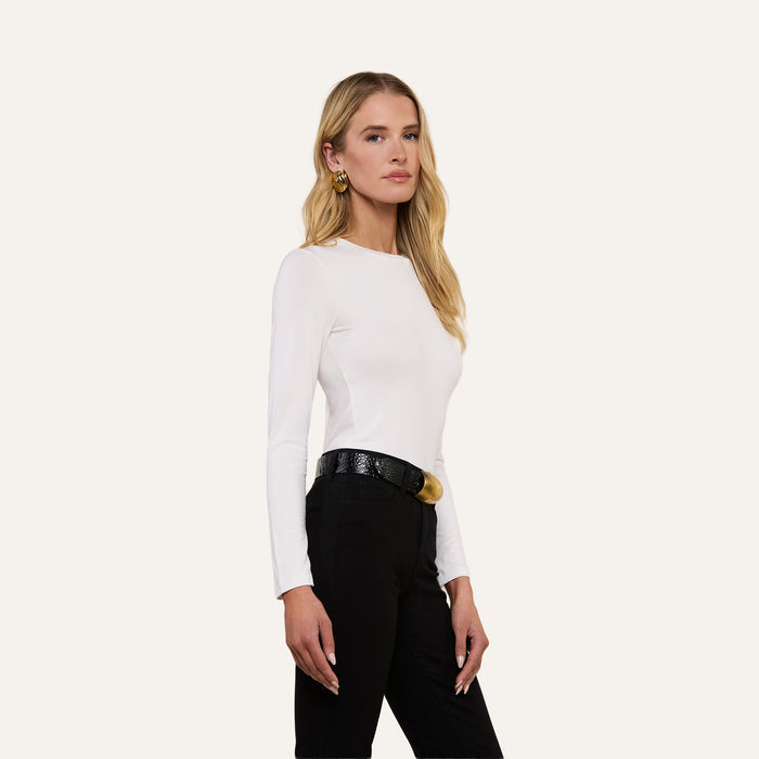 Wearing the Tess Long Sleeve Tee in white, a woman with long blonde hair poses against a plain light background. She pairs the slim-fit top with black pants, a textured black belt, and gold hoop earrings.