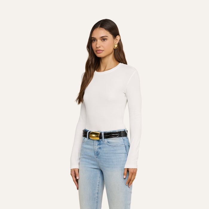 A woman with long brown hair wears the Tess Long Sleeve Tee—a slim-fit, ribbed micro modal white crew neck top—paired with light blue jeans, a black belt featuring a gold buckle, and large gold earrings. She stands against a plain light background, facing forward and slightly to the left.