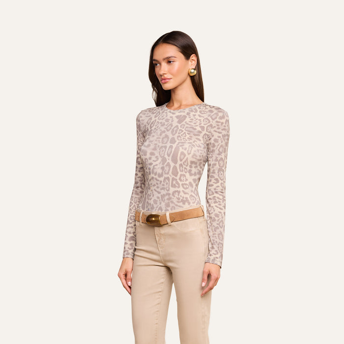A woman wearing the Tess Long Sleeve Tee in a slim fit grey leopard print, paired with tan pants, a brown belt, and gold earrings, stands against a plain light background.