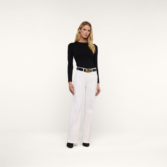 A woman stands against a plain white background, wearing the fitted black Tess Long Sleeve Tee, white high-waisted pants, a black belt with a gold buckle, and black shoes.
