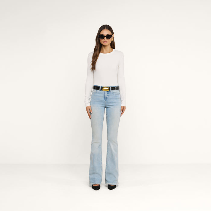 A woman wearing sunglasses, the Tess Long Sleeve Tee—a slim-fit white crew neck top made of ribbed micro modal—light blue flared jeans, a black belt, and black shoes stands against a plain white background.