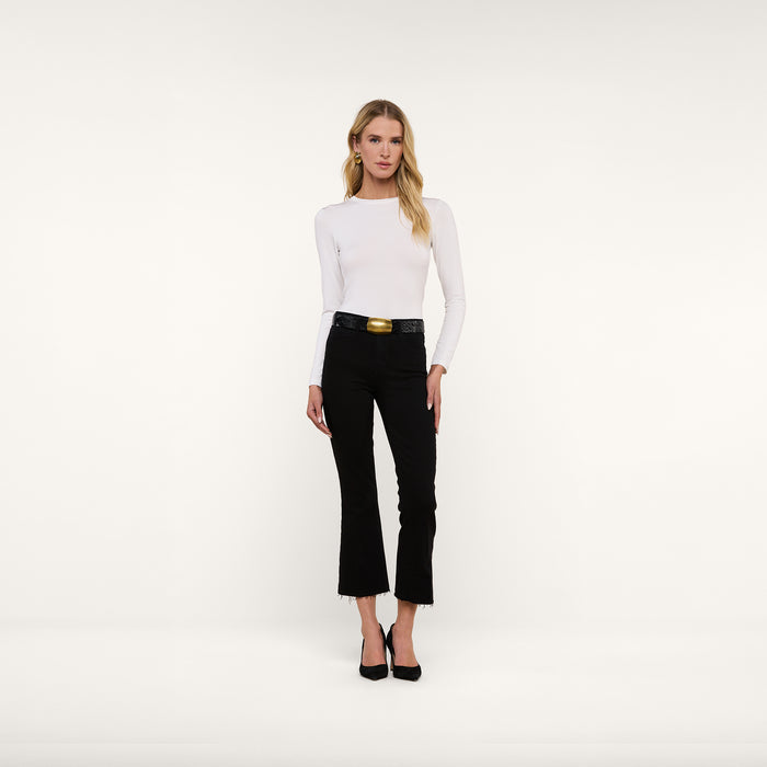 A woman stands facing forward against a plain white background, wearing the Tess Long Sleeve Tee—a slim-fit, microfiber jersey top in white—paired with black pants with a belt and black heels. The Tess Long Sleeve Tee offers a versatile layering option that adds a sleek touch to her outfit.