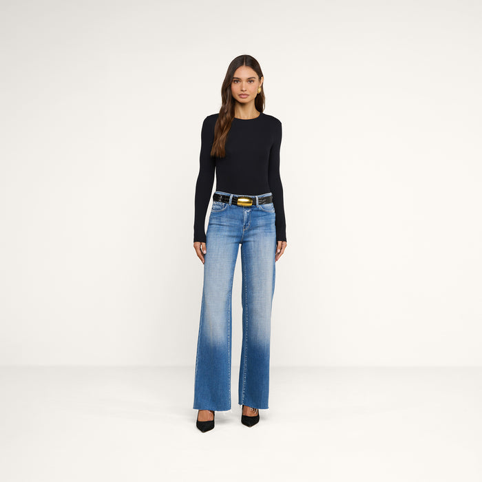 A woman stands against a plain white background, wearing the Tess Long Sleeve Tee in black, blue high-waisted jeans with a dark belt, and black pointed-toe heels. She has long brown hair and a neutral expression.