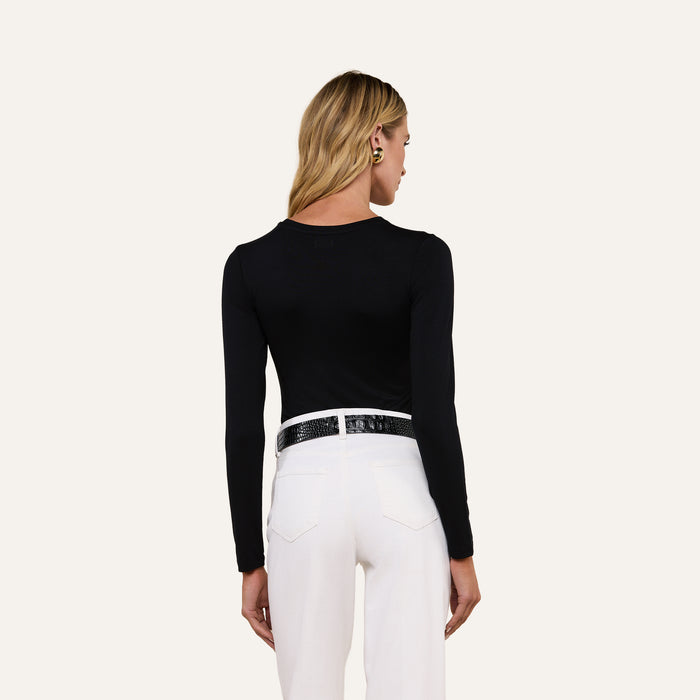 A person with long blonde hair is shown from the back, wearing the Tess Long Sleeve Tee in black, paired with white pants and a black belt, standing against a plain light background.
