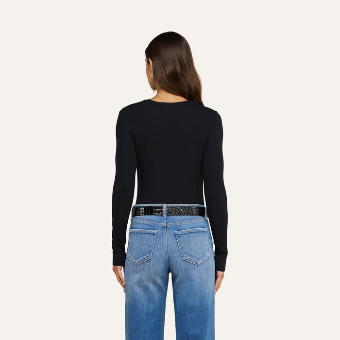 A woman with long brown hair, wearing the Tess Long Sleeve Tee in black and blue jeans, is seen from behind against a plain light background.